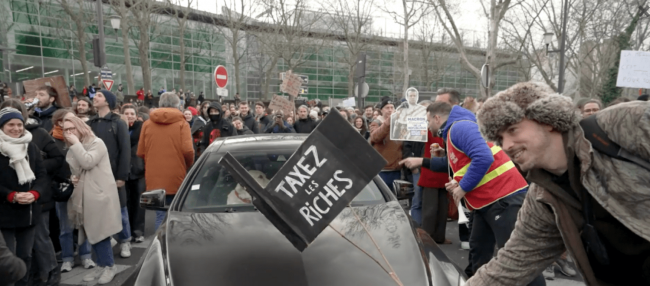 Photo: Still image of a French protester holding a sign over a vehicle. Screenshot/TRNN.